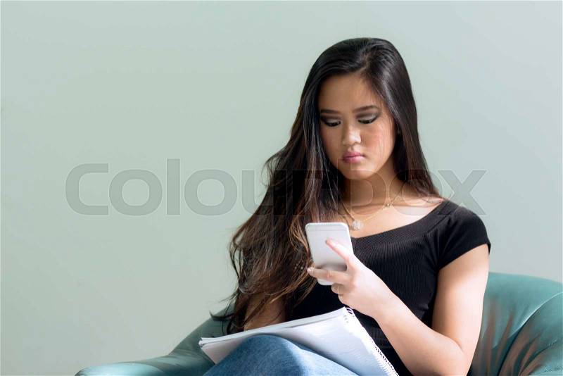 Asian woman with long black hair looking at smart phone while taking a break from studying her notebook, stock photo