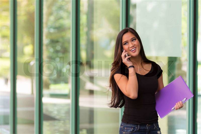 Smiling Asian woman talks on cell phone while walking next to glass building, stock photo