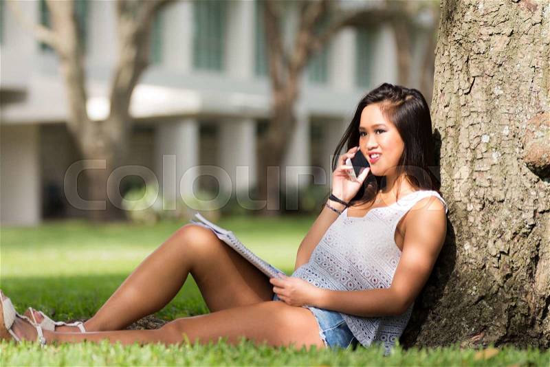 Smiling Asian girl talks on cell phone while studying under a tree on school campus, stock photo