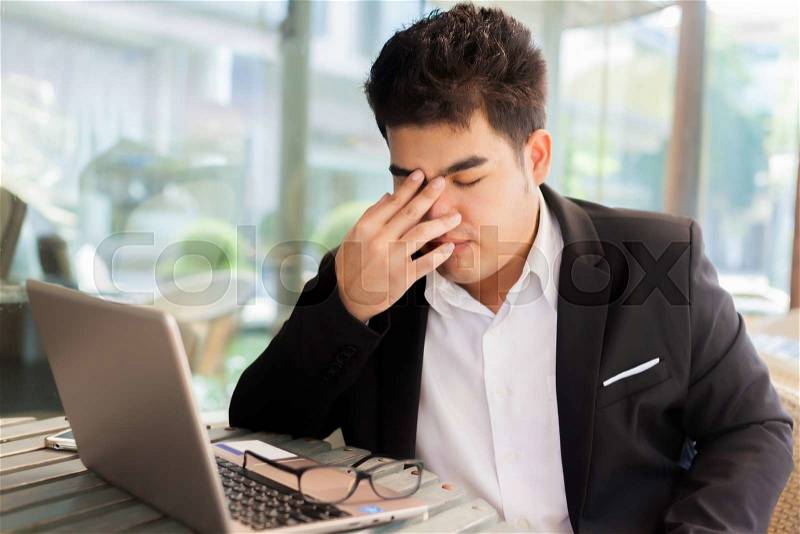 Young Asian businessman suffering from tired eyes after long hours of using laptop, stock photo