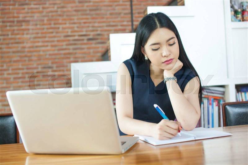 Beautiful Asian woman writing a notebook on table with laptop aside, stock photo