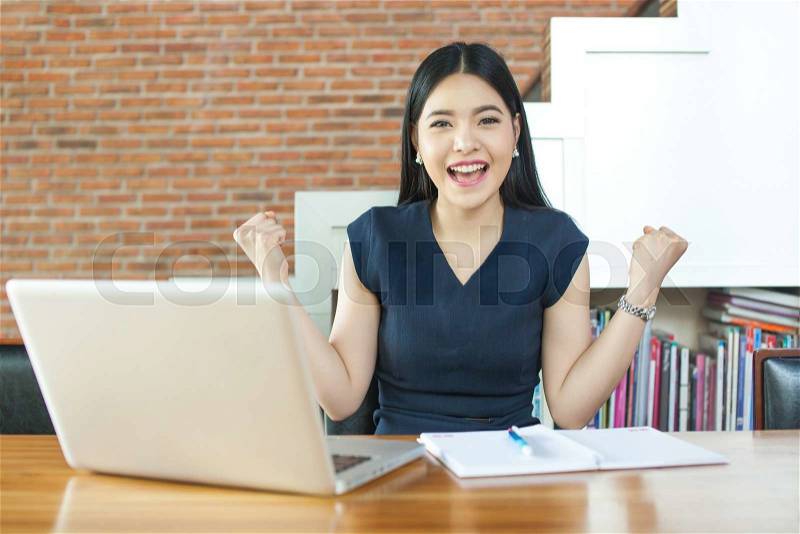 Excited Asian woman raising her arms while working on her laptop - success and business concept, stock photo