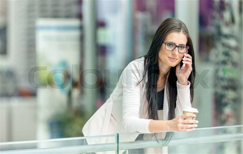 Portrait of young attractive business woman standing in the shopping mall with coffee and using her cell phone. Business break. Coffee break, stock photo