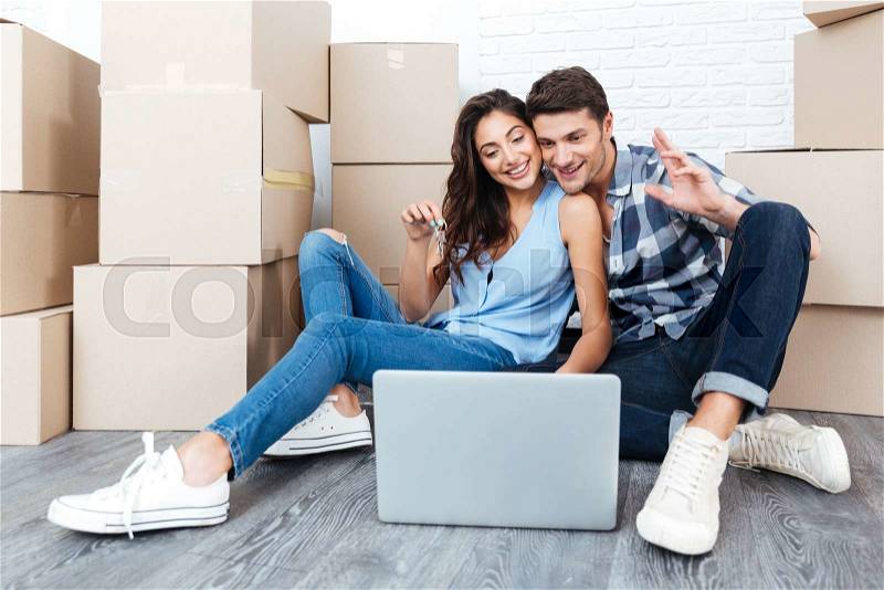 Young happy couple sitting on floor moving in a new house or apartment flat using computer laptop, stock photo