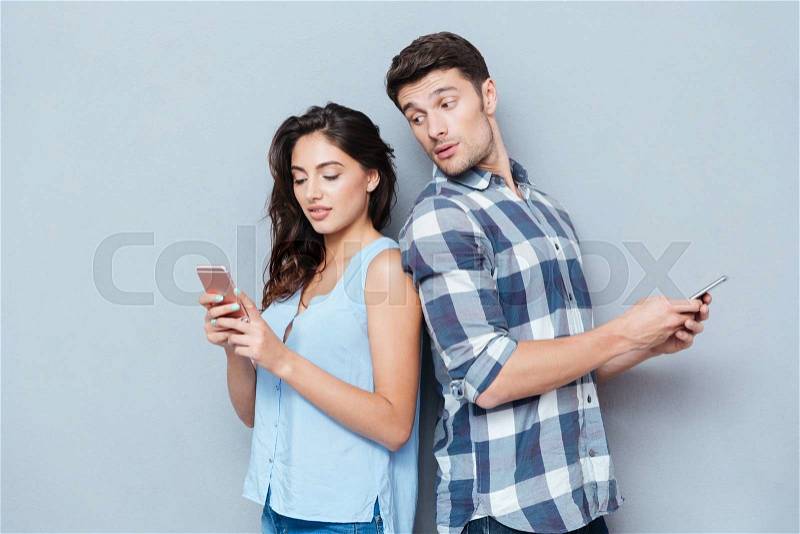 Jealous boyfriend watching his girlfriend texting on the phone isolated on gray background, stock photo