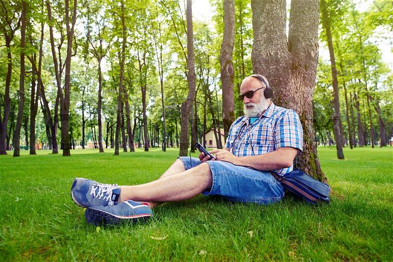 Aged man in modern fashionable clothes is sitting under the tree in picturesque park and listening to music on his smart phone, stock photo