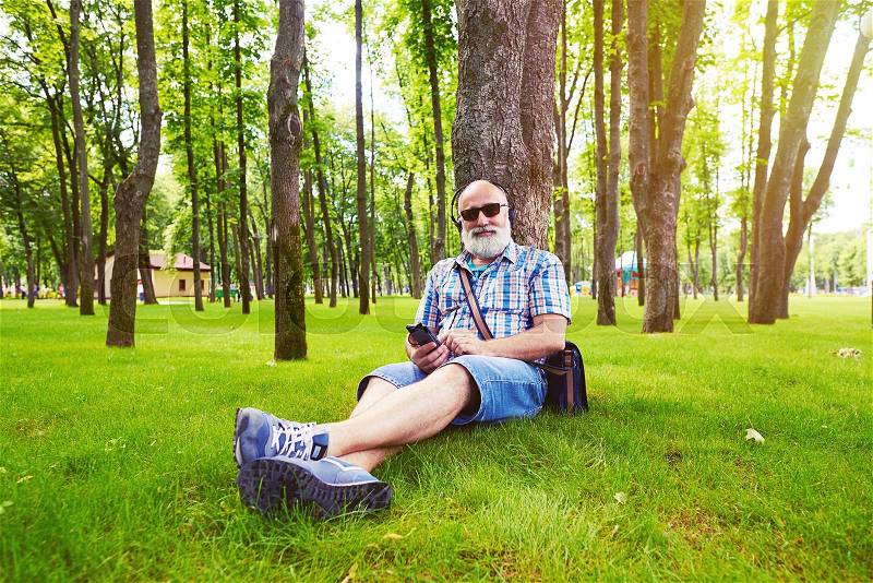 Aged stylish man is sitting under the tree on green grass and listening to music on his smart phone, stock photo