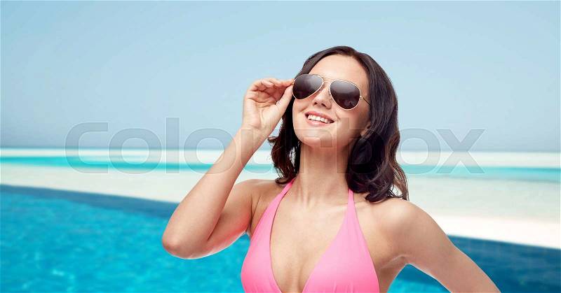 People, fashion, swimwear, summer and travel concept - happy young woman in sunglasses and pink bikini swimsuit over maldives beach with swimming pool background, stock photo