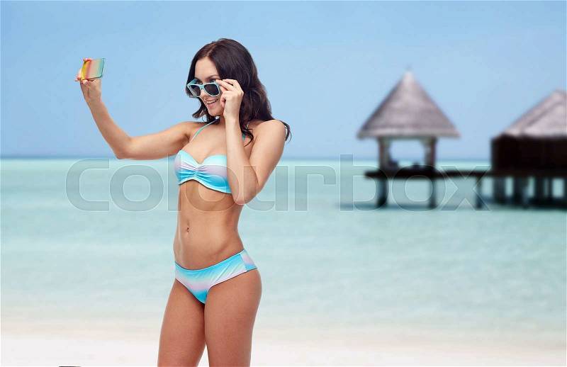 People, technology, summer and travel concept - happy young woman in bikini swimsuit and sunglasses taking selfie with smatphone over maldives beach with bungalow background, stock photo
