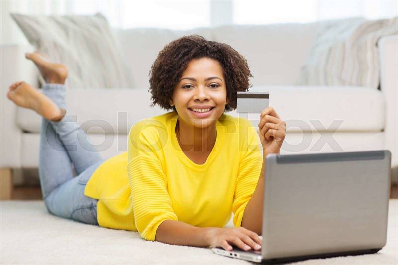 People, internet bank, online shopping, technology and e-money concept - happy african american young woman lying on floor with laptop computer and credit card at home, stock photo
