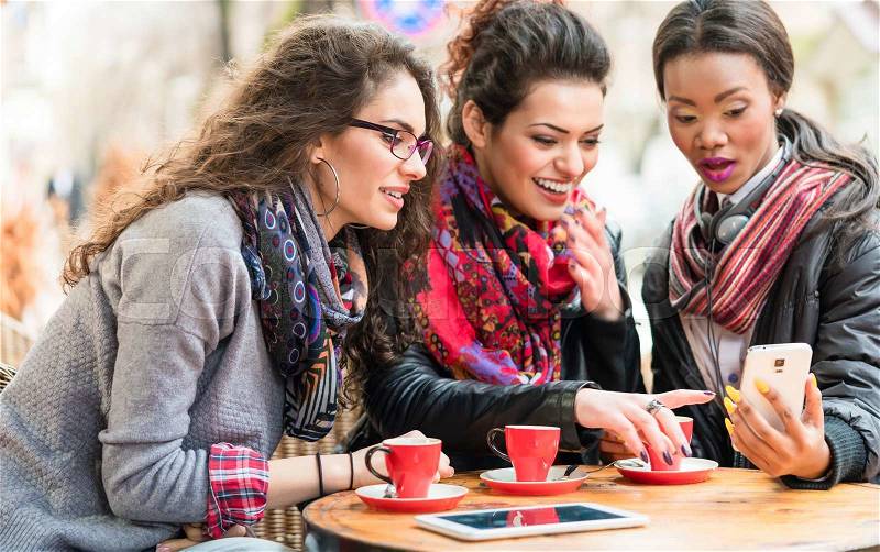 Multicultural group of women in cafe showing each other pictures on smart phone and chatting, stock photo