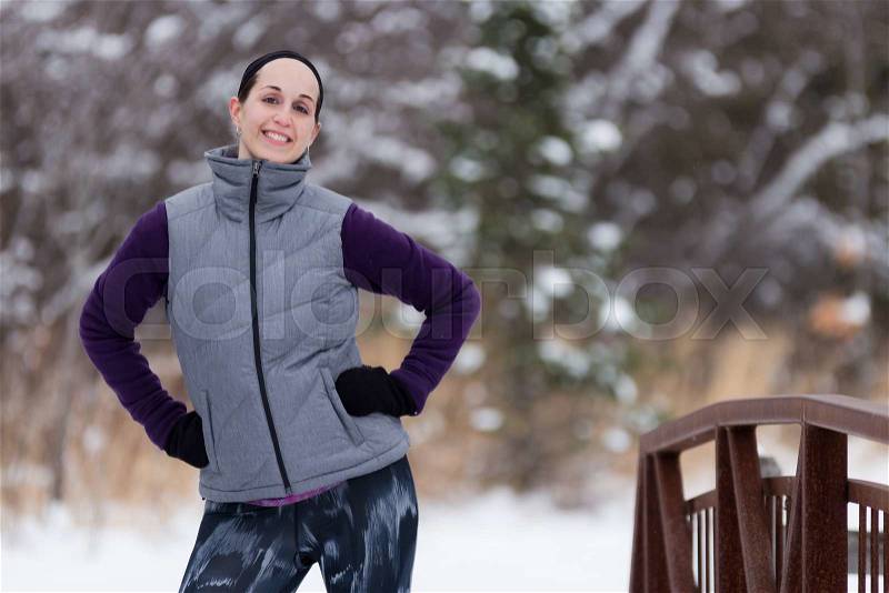 Fit woman in cold weather workout attire near forest during winter smiles at camera. Fitness and wellness lifestyle concept, stock photo