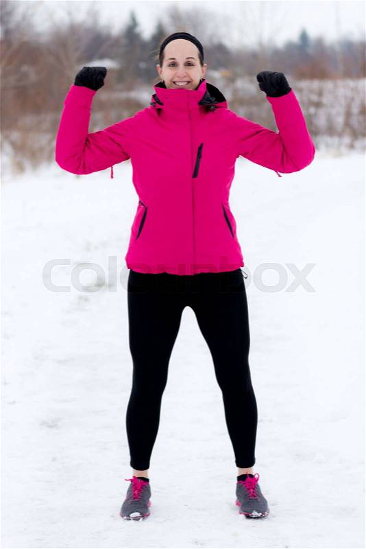 Smiling fit woman in cold weather workout attire near forest during winter flexing arms . Fitness and wellness lifestyle concept, stock photo