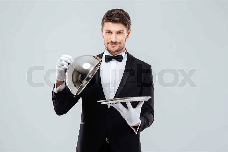 Happy young waiter in tuxedo and bowtie lifting metal cloche from serving tray, stock photo