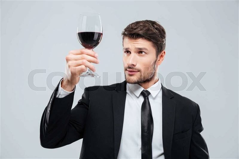 Serious young man sommelier in suite tasting red wine in glass over white background, stock photo