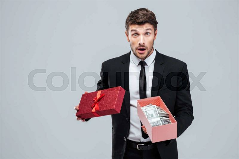 Surprised young businessman holding opened gift box with money over white background, stock photo