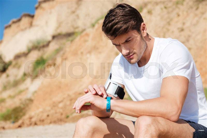 Concentrated young sportsman sitting and using fitness tracker on the beach, stock photo