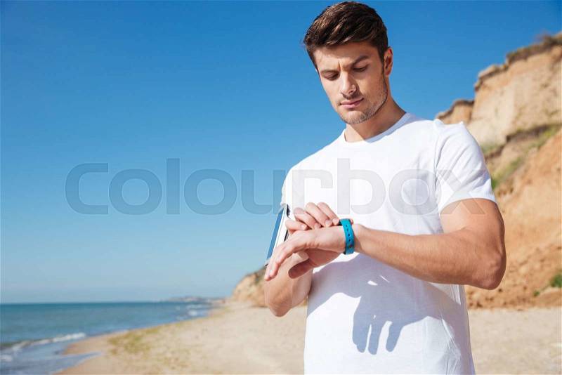 Handsome young sportsman standing and using smart watch on the beach, stock photo