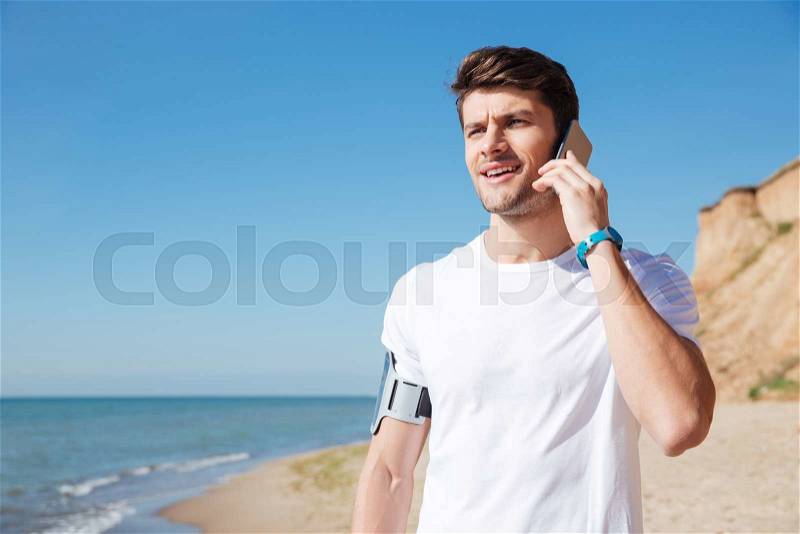 Happy young sportsman talking on mobile phone on the beach, stock photo