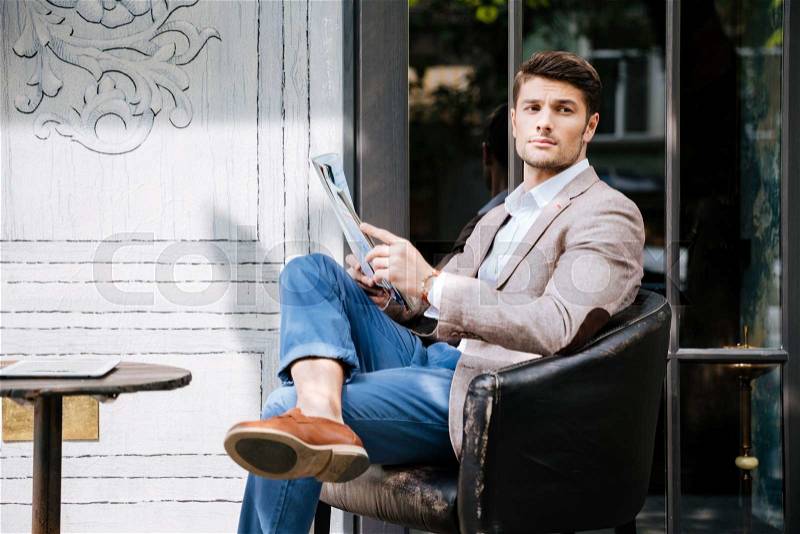 Pensive young man sitting and reading magazine in outdoor cafe, stock photo