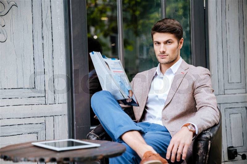 Serious young man in jeans and jacket reading magazine in outdoor cafe, stock photo