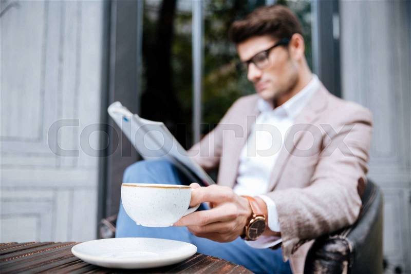 Thoughtful young man in glasses with cup of coffee sitting and reading magazine in outdoor cafe, stock photo