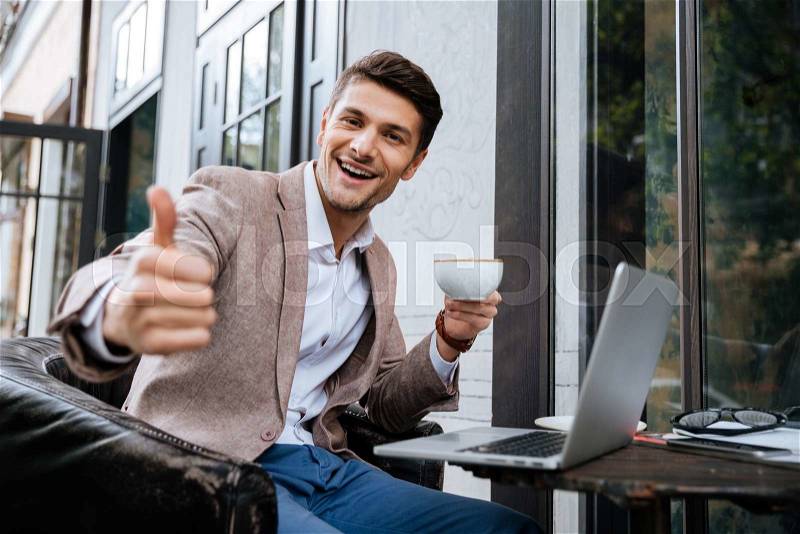Cheerful young man with laptop showing thumbs up and drinking coffee in outdoor cafe, stock photo