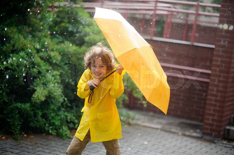 Boy of 8-9 years in the rain. It holds a yellow umbrella. The child with a smile looks in the camera having squinted. It has wild curly hair and a nice face. The boy is dressed in yellow raincoat, stock photo