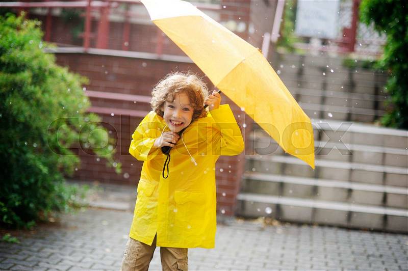 The little fellow in a bright yellow raincoat is in the yard under a pouring rain. Two hands he holds an open yellow umbrella. Large drops of a rain. To the boy it is very cheerful. He with a smile looks in the camera, stock photo
