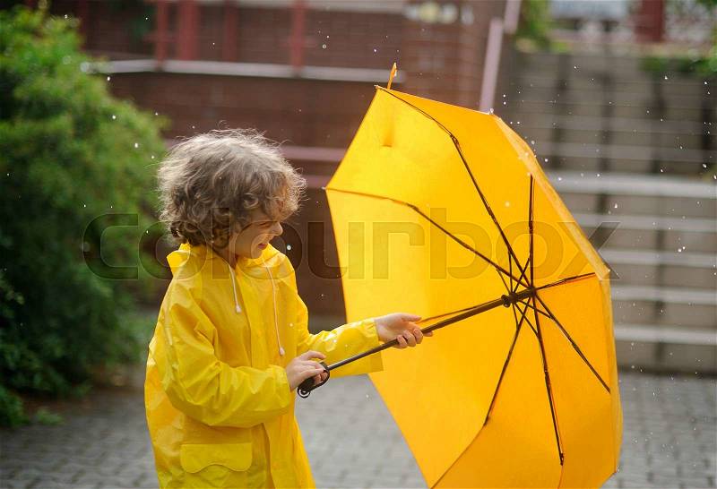 The boy in a bright yellow raincoat opens an umbrella during a rain. The child is in the yard of the house. Drops of a rain fall to it on the head. The boy with a cheerful smile opens a yellow umbrella, stock photo