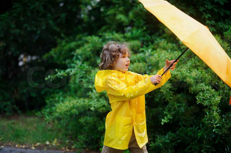 Curly boy in a bright yellow raincoat with an umbrella against a magnificent green bush, stock photo