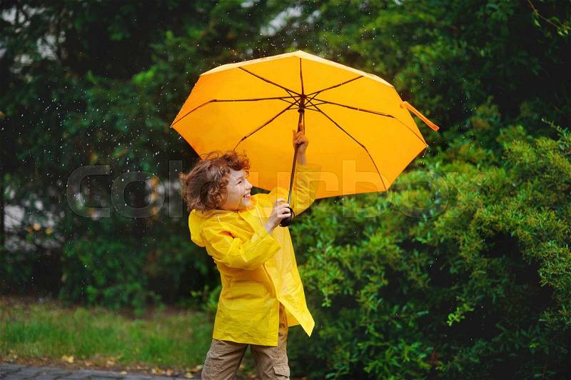 Boy of 8-9 years stand in the rain with a big yellow umbrella. He likes such weather. He cheerfully laughs. Behind the back of the child a magnificent green bush, stock photo
