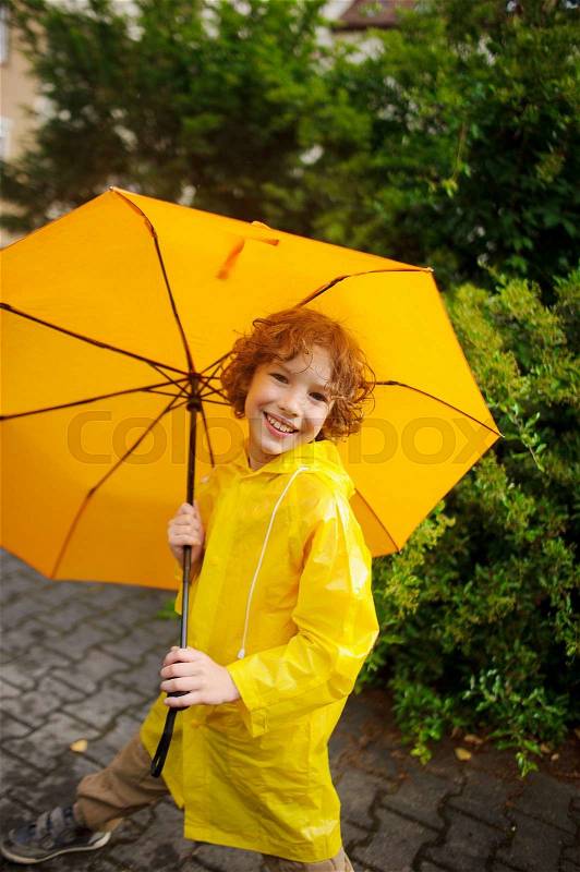 The cute boy of 8-9 years under a yellow umbrella. The boy in a bright yellow raincoat holds a big umbrella in hand. The child looks in the camera with a smile. Behind magnificent wet bush, stock photo