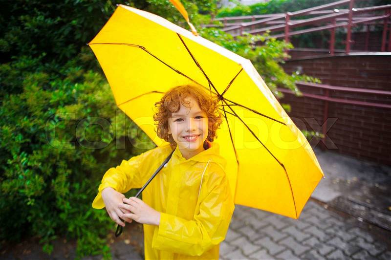 The nice fellow of 8-9 years under a yellow umbrella. The boy in a bright yellow raincoat holds a big umbrella in hand. The child looks in the camera with a smile. It likes to walk under a drizzle on the wet sidewalk, stock photo