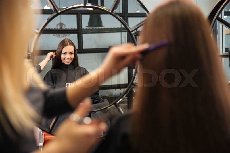 Mirror reflection of young woman getting her hairdo by stylist at parlor, stock photo