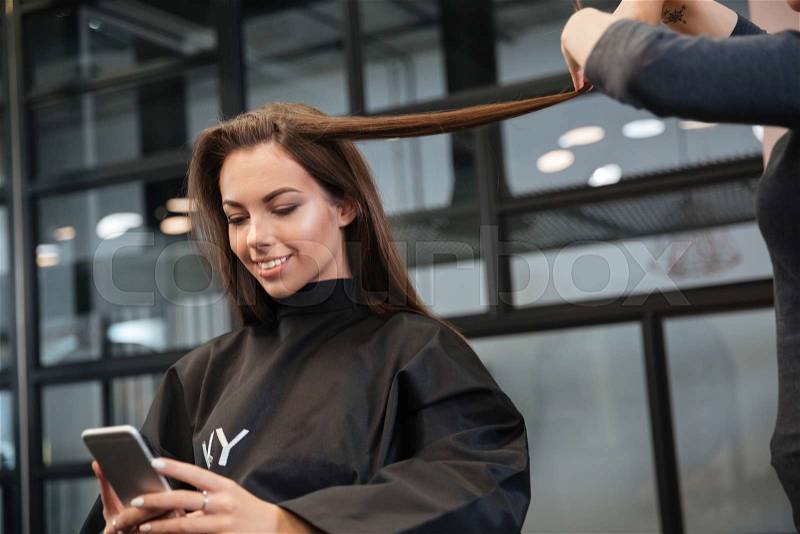 Happy young woman with smartphone and hairdresser making hair styling at salon, stock photo
