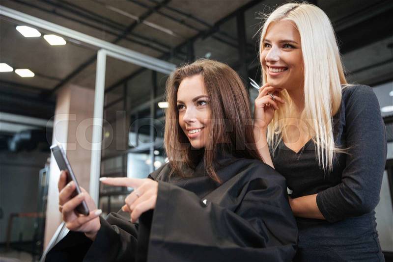 Happy young woman pointing at smartphone and hairdresser making hair styling at salon, stock photo