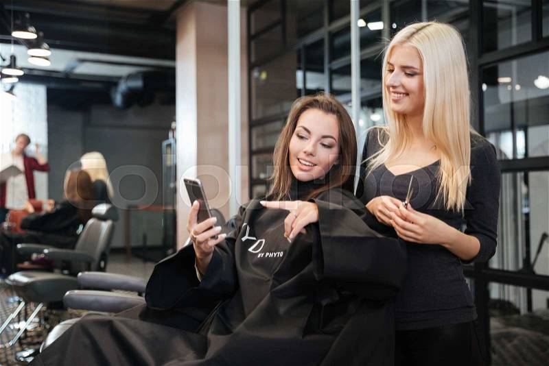Happy young woman pointing at smartphone and hairdresser making hair styling at salon, stock photo
