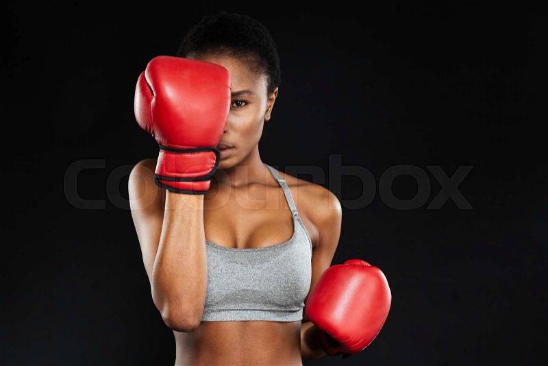 Strong intense young fitness woman covered her face with boxing gloves over black background, stock photo