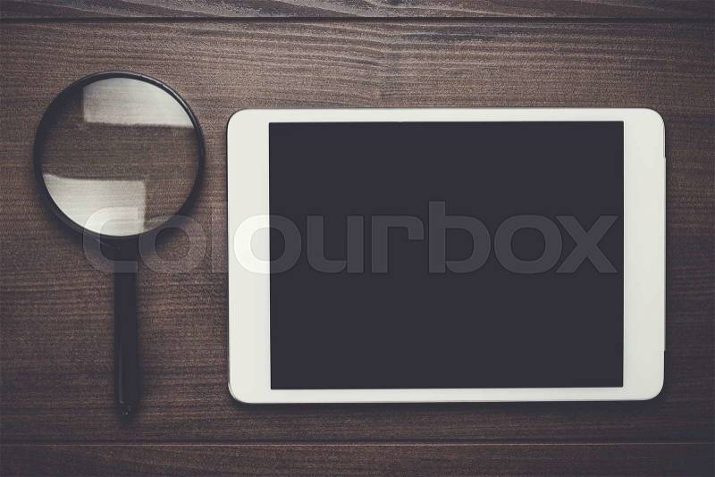 Magnifying glass and tablet computer on brown wooden background, stock photo