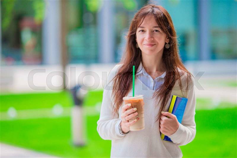 Happy young student girl with a coffee-to-go, walking in a summer park and holding books for reading, stock photo