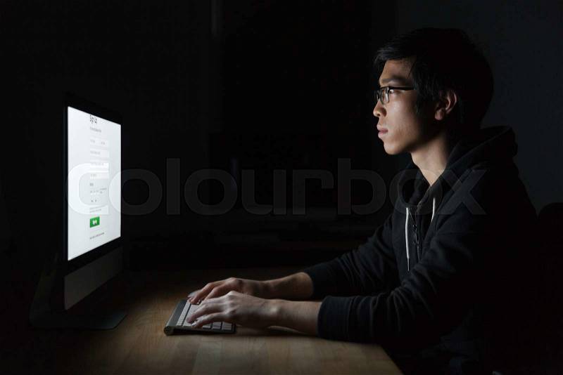 Concentrated asian young man in glasses using computer in the evening in office, stock photo