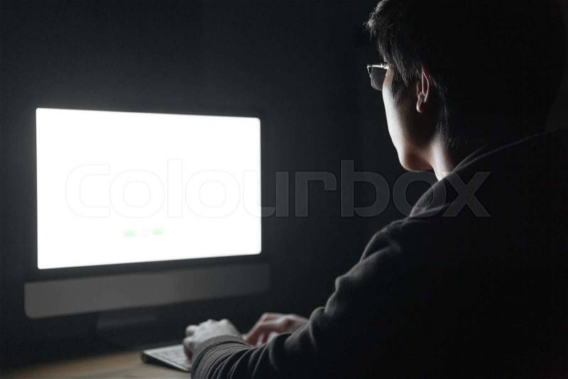 Back view of young man in glasses using blank screen computer in dark room, stock photo