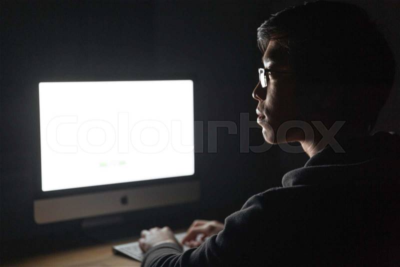 Profile of focused young man sitting and using blank screen computer in dark room, stock photo
