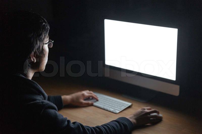 Concentrated young man in glasses sitting at the table and using computer in dark room, stock photo