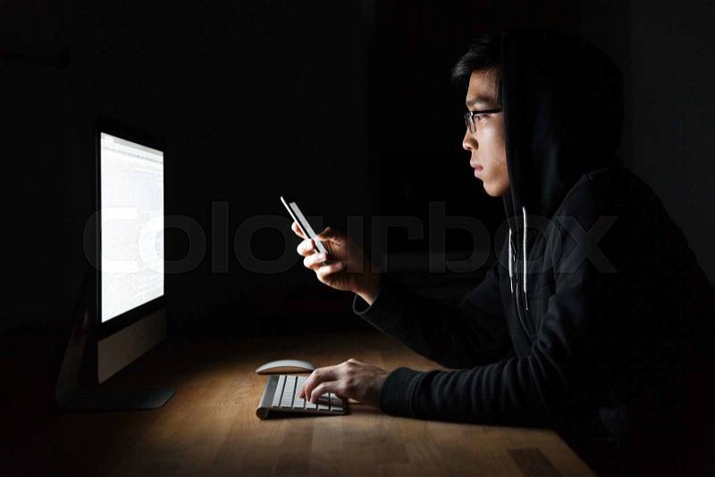 Serious young hacker using laptop and mobile phone in dark room, stock photo