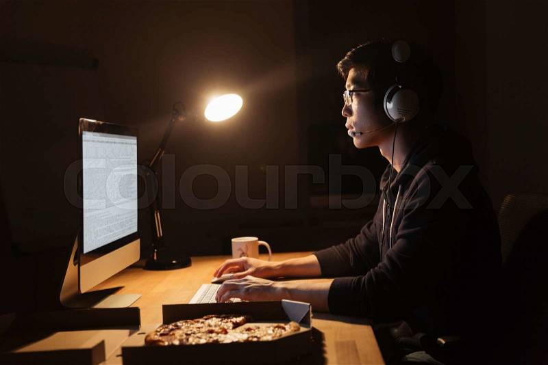 Focused asian man in headphones working with computer and eating pizza at the table in dark room, stock photo