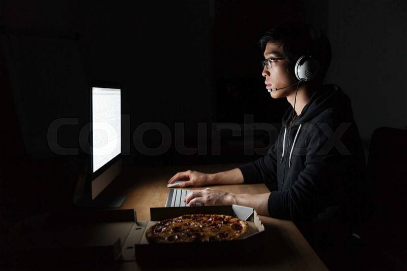 Man in headphones working with computer and eating pizza in dark room, stock photo