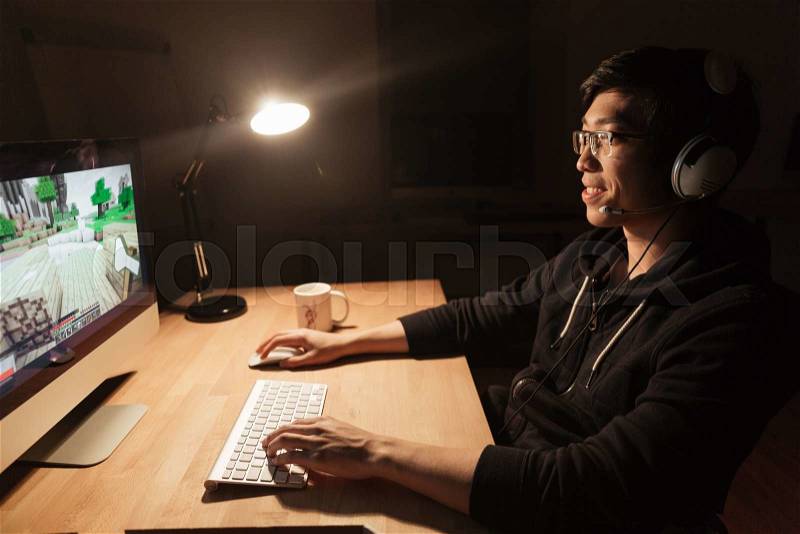 Smiling asian young male gamer playing computer game in dark office, stock photo