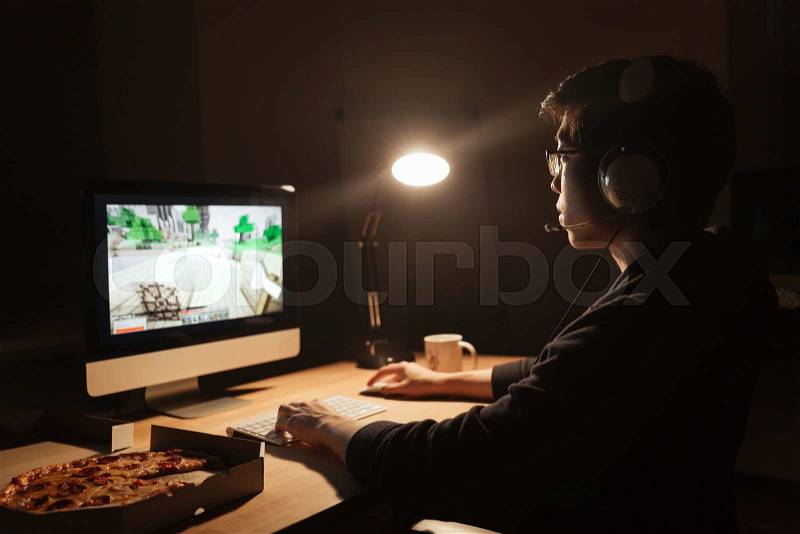 Male gamer playing computer game and eating pizza in dark room, stock photo